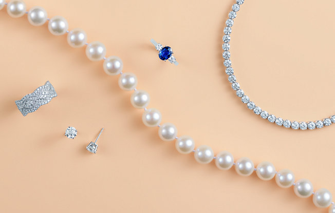 A pearl necklace, diamond ring, diamond stud earrings, sapphire engagement ring and diamond tennis bracelet on an orange background