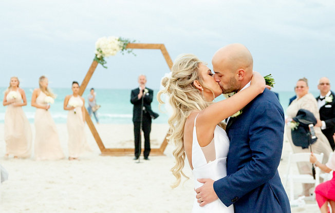 A bride and groom kiss in front of an arch on the beach at The National Hotel