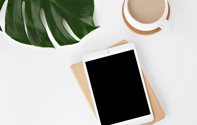 An Ipad, a full cup of coffee and a plam leaf sit on a clean white table