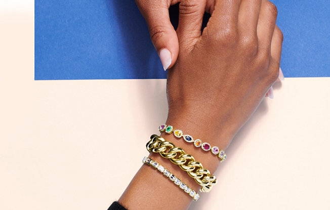 A woman wearing a chunky gold bracelet, a multicolored gemstone bracelet and a diamond tennis bracelet on her wrist