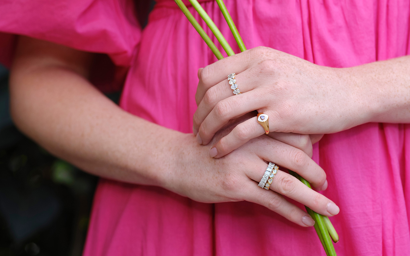 Photo of woman’s hands, she is wearing multiple diamond rings and a pink dress. 