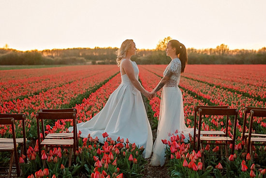 'two women in wedding dresses holding hands in a field of orange flowers