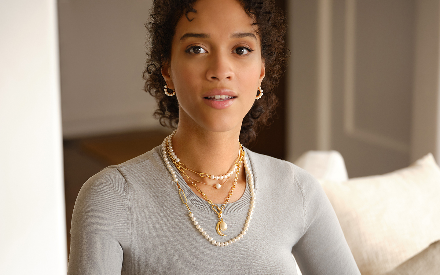 Woman wearing multiple pearl and gemstone jewels including 3 necklaces and a set of pearl hoop earrings. 