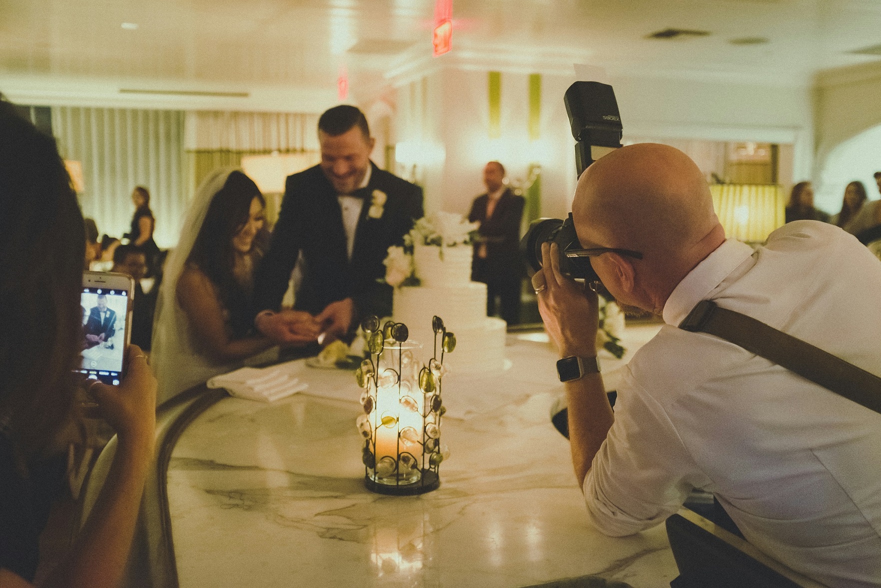 Bride and groom cutting their cake as they’re photographed with DSLRs and a camera phone.