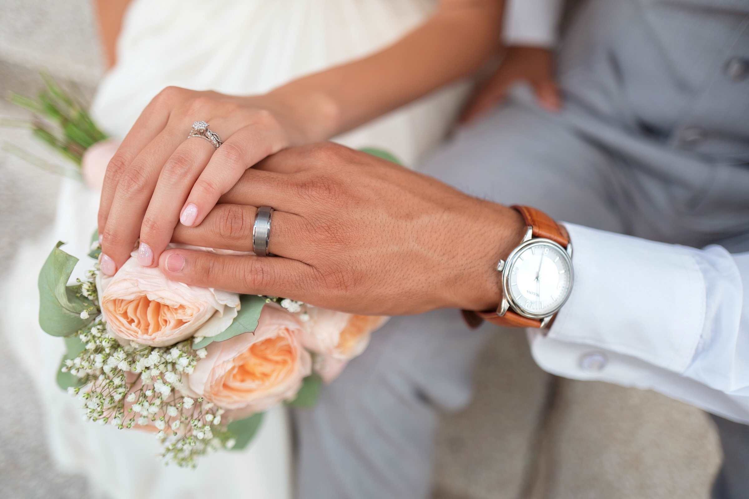 Photo of a bride and groom with their hands on the bride’s bouquet. 