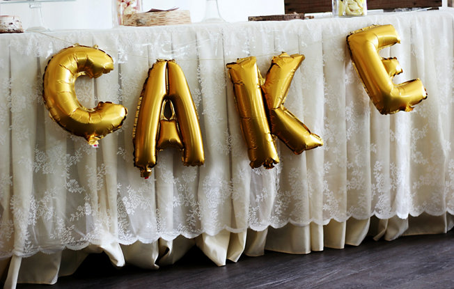Gold letter balloons that spell out the word cake attached to a table dresses with a lace tablecloth