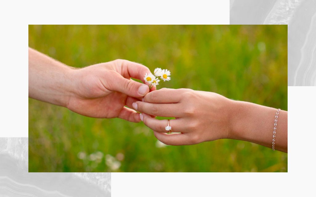 Hands of an engaged couple holding small wildflowers, with the engagement ring prominently displayed. 