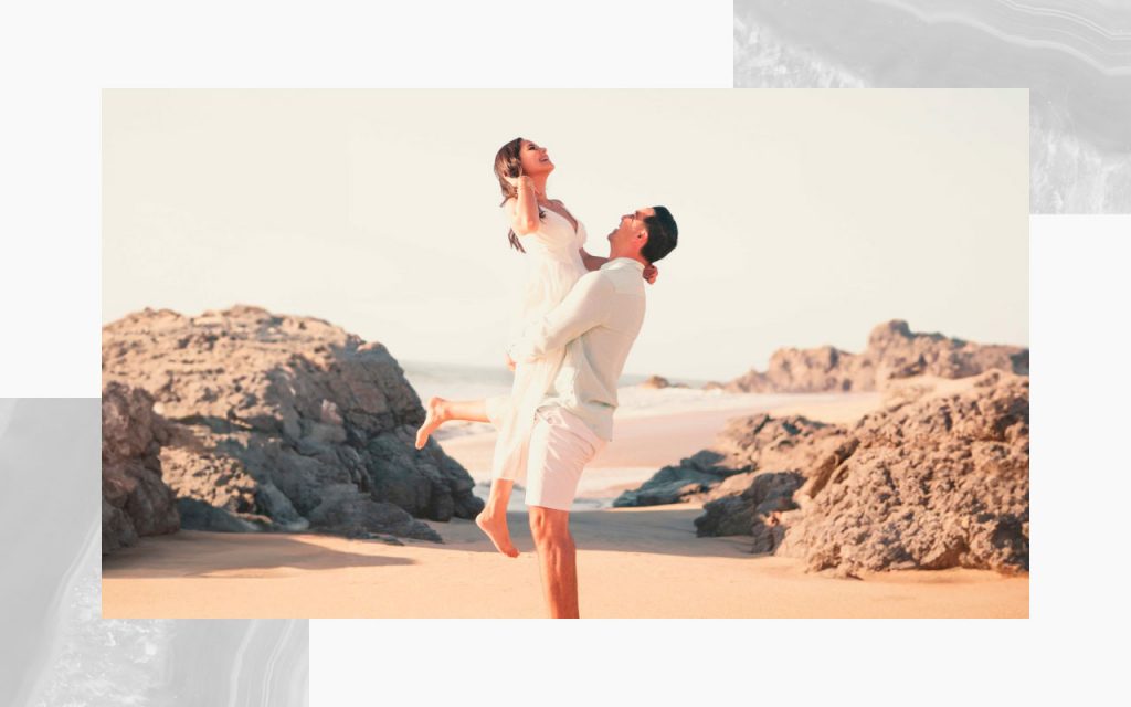 An engaged couple at the beach, with one lifting the other up.