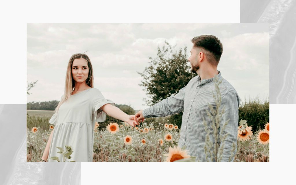An engaged couple reaching hands out towards each other in a field of sunflowers. 