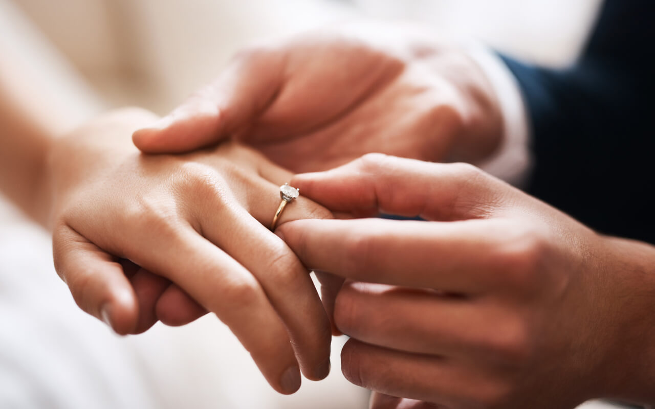 Close-up of someone slipping a gold solitaire onto their partner’s hand, showing a classic moment tied to engagement ring trends