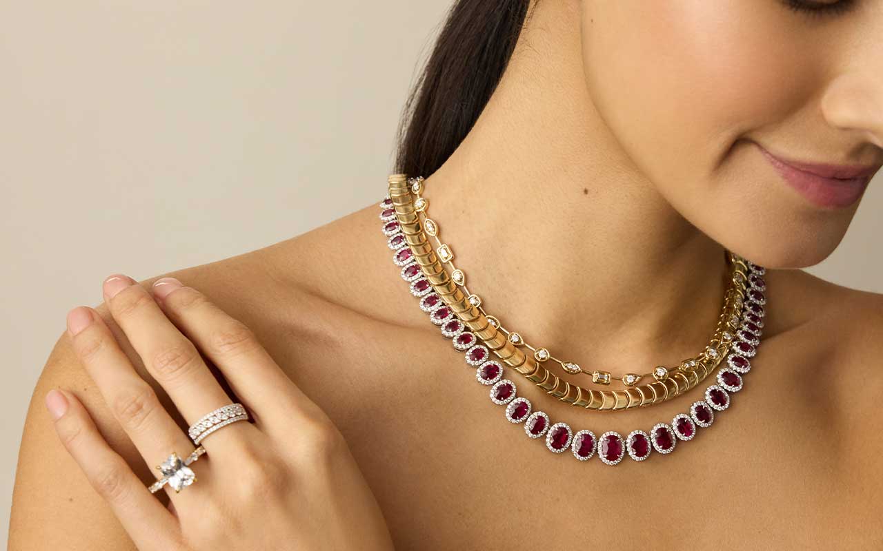 A model in a red dress smiles while showing off rings, necklaces, and earrings from the Blue Nile Modern Vintage Romance collection.
