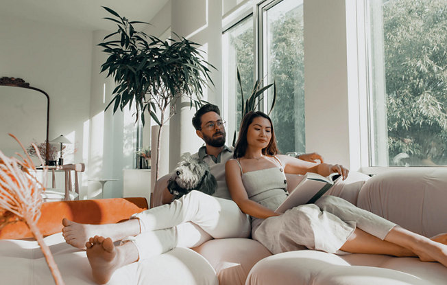 A couple sits on a beige couch with their dog while the woman reads a book