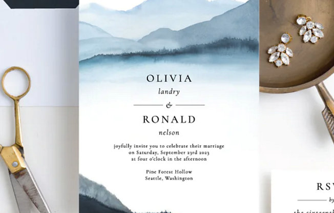 A Paper Culture wedding invitation with mountain design on a white table next to diamond earrings and scissors