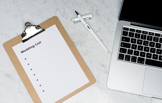 A wedding checklist, a pen and laptop sit on a marble table